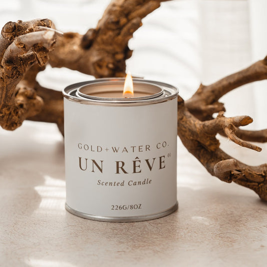 A lit scented candle from GOLD+WATER.CO UN RÈVE in a metal tin with a gray label, placed on a surface with a piece of driftwood.